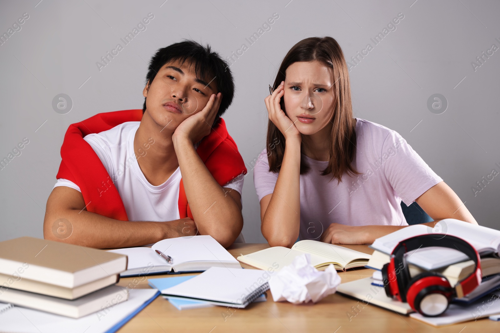 Photo of Tired students with books preparing for exam at table near grey wall