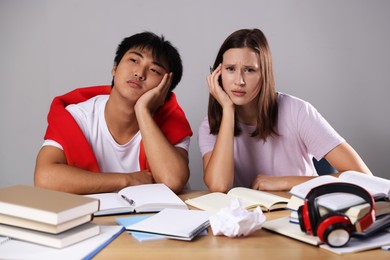 Photo of Tired students with books preparing for exam at table near grey wall
