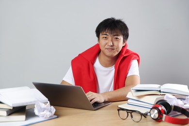 Photo of Student with laptop and books preparing for exam at wooden table near grey wall