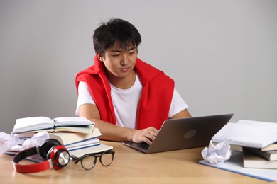 Photo of Student with laptop and books preparing for exam at wooden table near grey wall