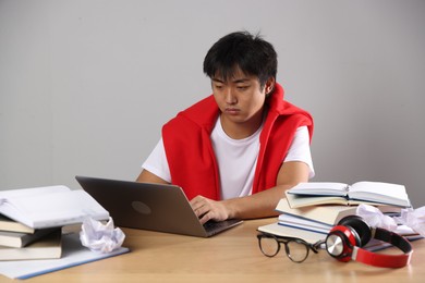 Photo of Student with laptop and books preparing for exam at wooden table near grey wall