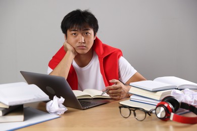 Photo of Student with laptop and books preparing for exam at wooden table near grey wall