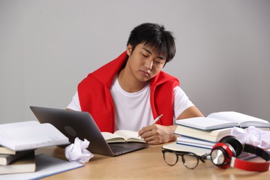 Photo of Student with laptop and books preparing for exam at wooden table near grey wall
