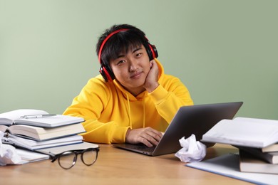 Photo of Student in headphones with laptop and books preparing for exam at wooden table near green wall