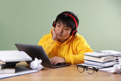 Photo of Student in headphones with laptop and books preparing for exam at wooden table near green wall