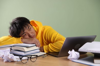 Photo of Tired student with laptop and books before exam at wooden table near green wall