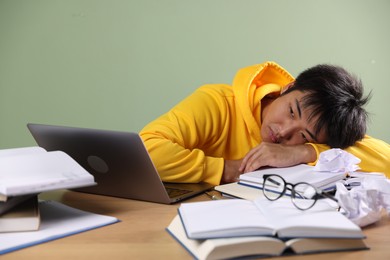Photo of Tired student with laptop and books before exam at wooden table near green wall