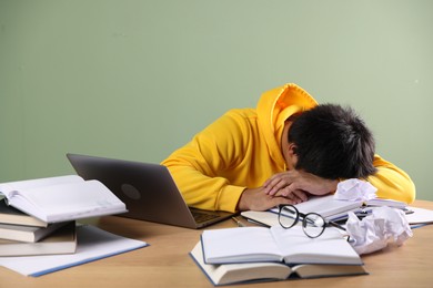 Photo of Tired student with laptop and books before exam at wooden table near green wall