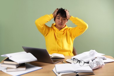 Photo of Tired student with laptop and books before exam at wooden table near green wall
