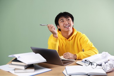 Photo of Happy student with laptop and books preparing for exam at wooden table near green wall