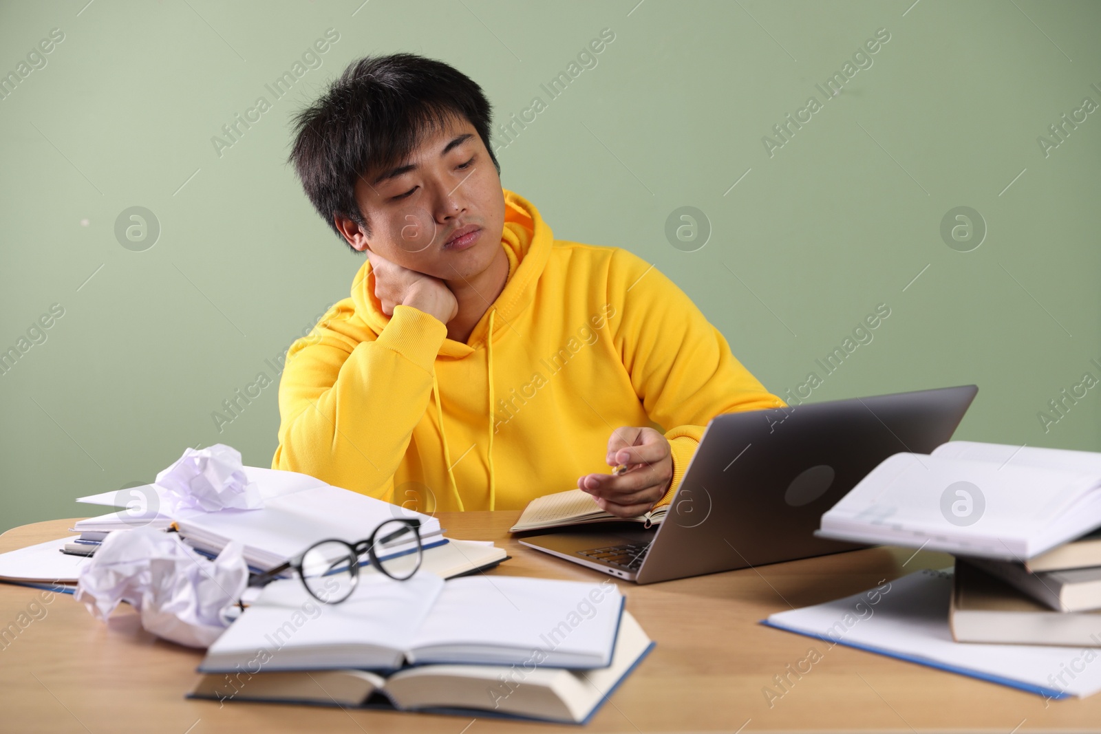 Photo of Student with laptop and books preparing for exam at wooden table near green wall