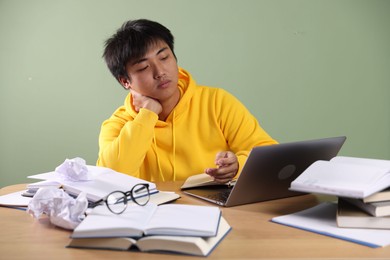 Photo of Student with laptop and books preparing for exam at wooden table near green wall