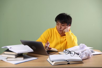 Photo of Student with laptop and books preparing for exam at wooden table near green wall