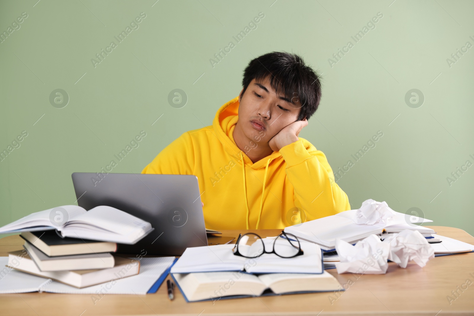 Photo of Student with laptop and books preparing for exam at wooden table near green wall