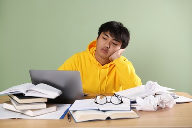 Photo of Student with laptop and books preparing for exam at wooden table near green wall