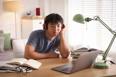 Photo of Student in headphones with laptop and books preparing for exam at table indoors