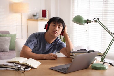 Photo of Student in headphones with laptop and books preparing for exam at table indoors