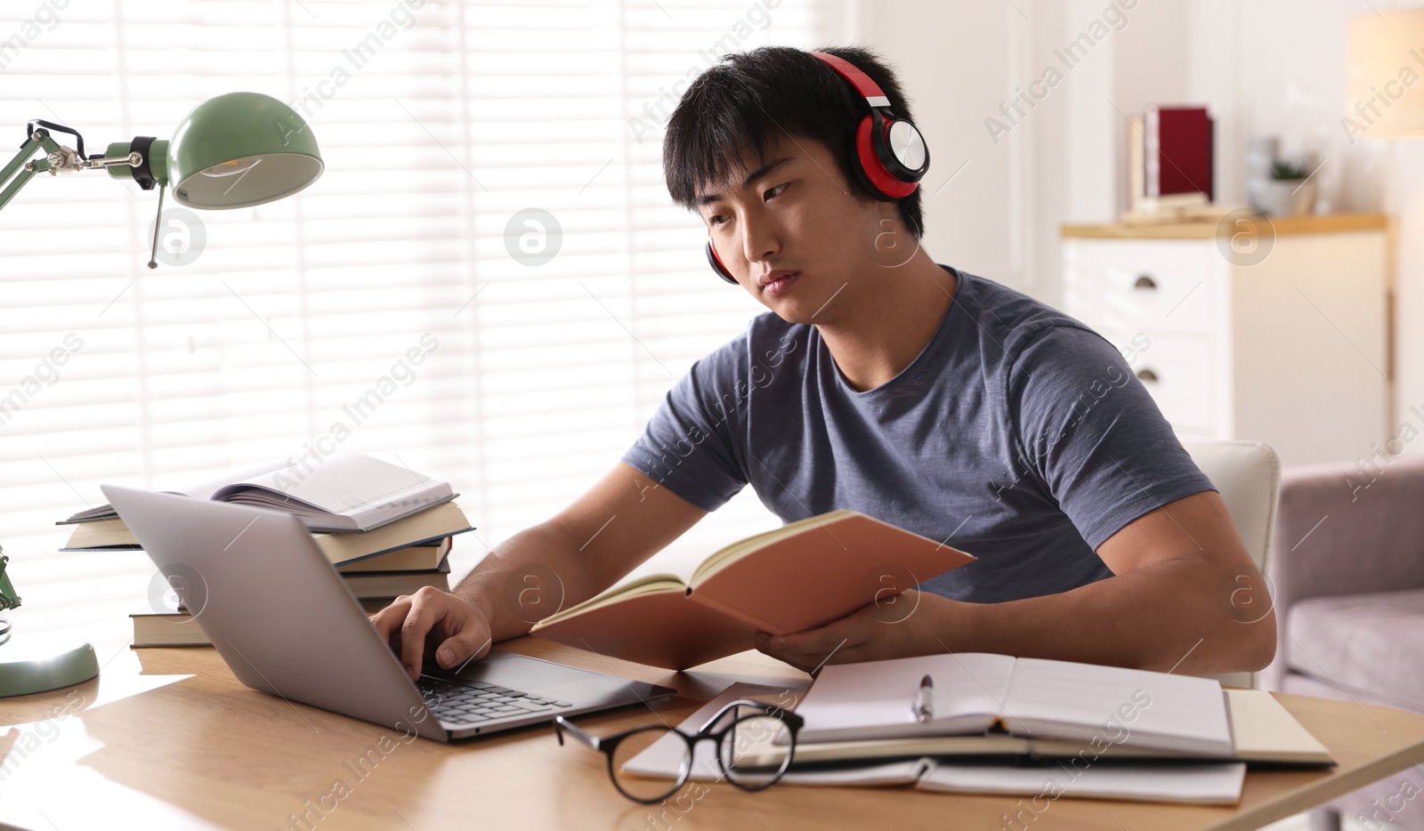 Photo of Student in headphones with laptop and books preparing for exam at table indoors