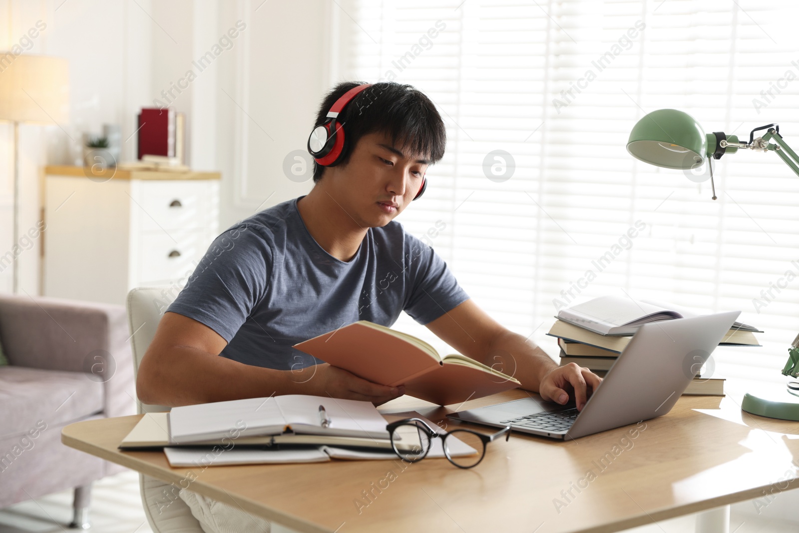 Photo of Student in headphones with laptop and books preparing for exam at table indoors