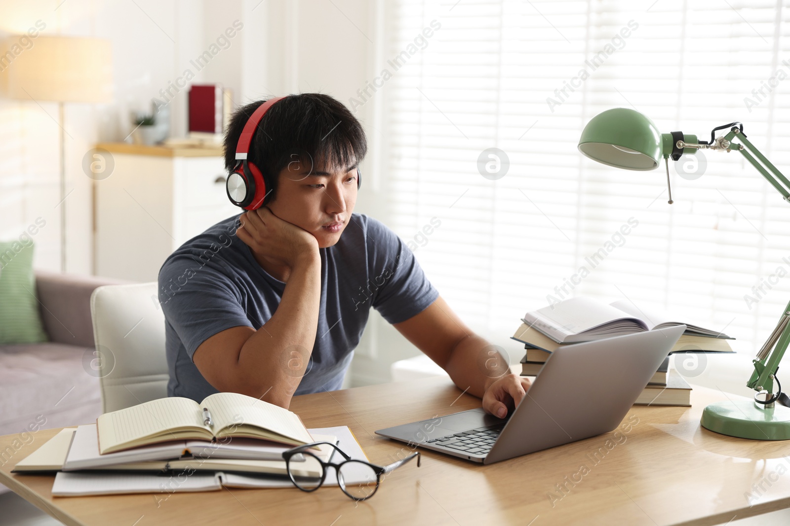 Photo of Student in headphones with laptop and books preparing for exam at table indoors