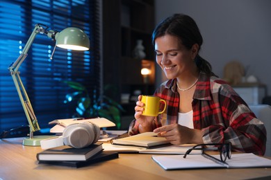 Photo of Happy student with cup of drink preparing for exam at wooden desk indoors