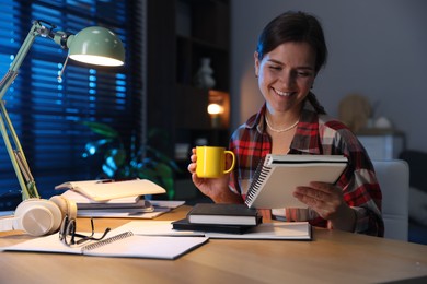 Photo of Happy student with cup of drink preparing for exam at wooden desk indoors