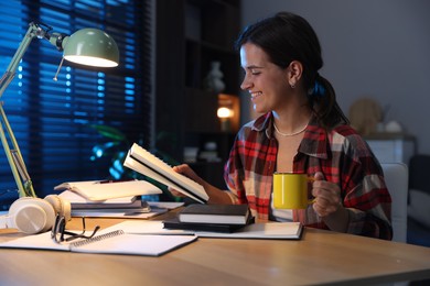 Photo of Happy student with cup of drink preparing for exam at wooden desk indoors