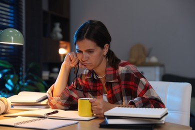 Photo of Tired student with cup of drink preparing for exam at wooden desk indoors