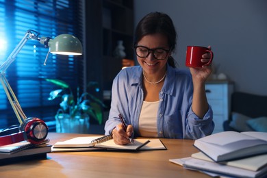 Photo of Happy student with cup of drink preparing for exam at wooden desk indoors