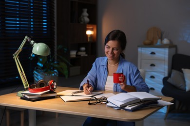 Photo of Happy student with cup of drink preparing for exam at wooden desk indoors