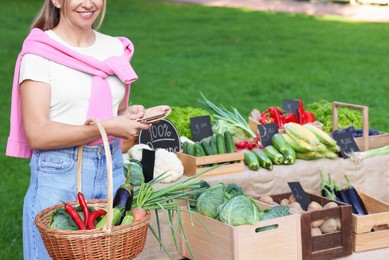 Photo of Woman with different fresh vegetables in wicker basket at farmer's market, closeup