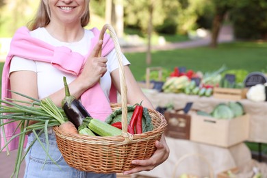 Photo of Woman with different fresh vegetables in wicker basket at farmer's market, closeup