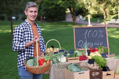 Photo of Happy man with different vegetables in wicker basket at farmer's market