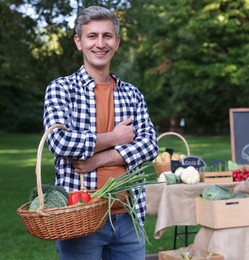 Photo of Happy man with different vegetables in wicker basket at farmer's market