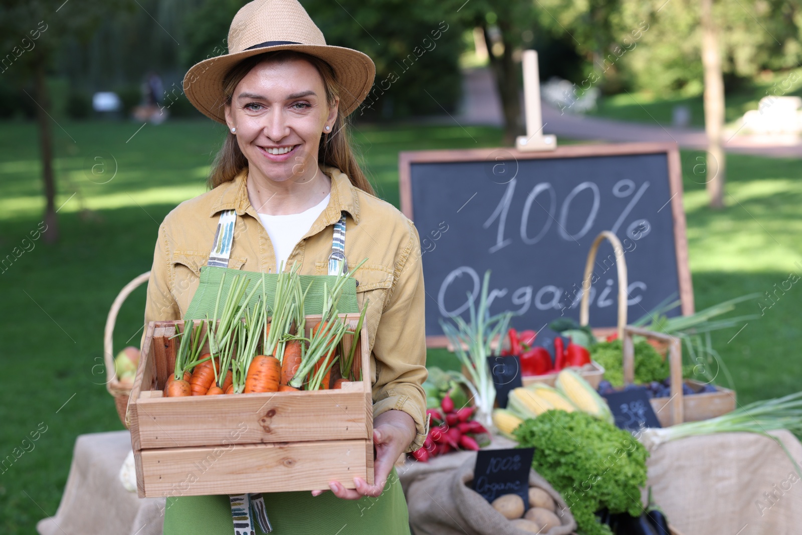 Photo of Happy farmer with crate of carrots near stall with different fresh vegetables at market
