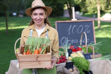 Photo of Happy farmer with crate of carrots near stall with different fresh vegetables at market