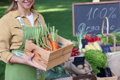 Photo of Farmer with crate of carrots near stall with different fresh vegetables at market, closeup