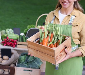 Photo of Farmer with crate of carrots near stall with different fresh vegetables at market, closeup