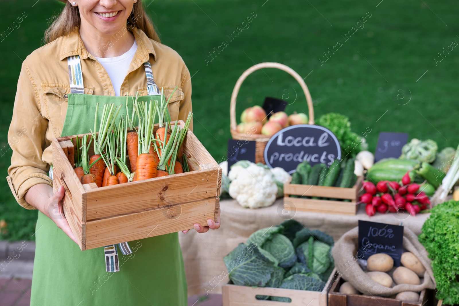Photo of Farmer with crate of carrots near stall with different fresh vegetables at market, closeup