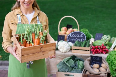 Photo of Farmer with crate of carrots near stall with different fresh vegetables at market, closeup