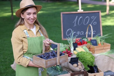 Photo of Happy farmer with basket of plums near stall with different fresh vegetables at market