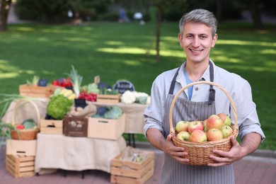 Photo of Happy farmer with apples in wicker basket near stall at market
