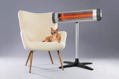 Photo of Cute little dog on armchair near electric heater against grey background