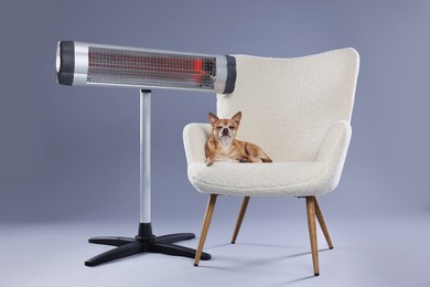 Photo of Cute little dog on armchair near electric heater against grey background