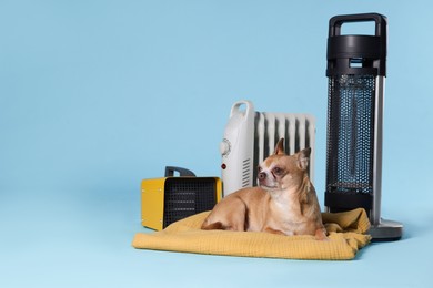 Photo of Cute little dog on blanket near electric heaters against light blue background, space for text