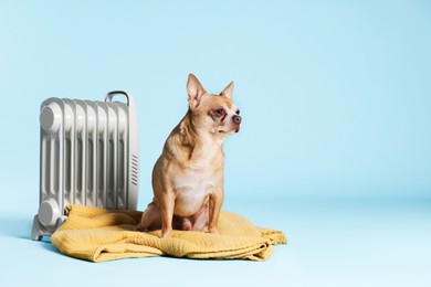 Photo of Cute little dog on blanket near electric heater against light blue background, space for text