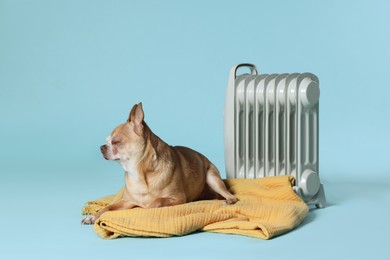 Photo of Cute little dog on blanket near electric heater against light blue background