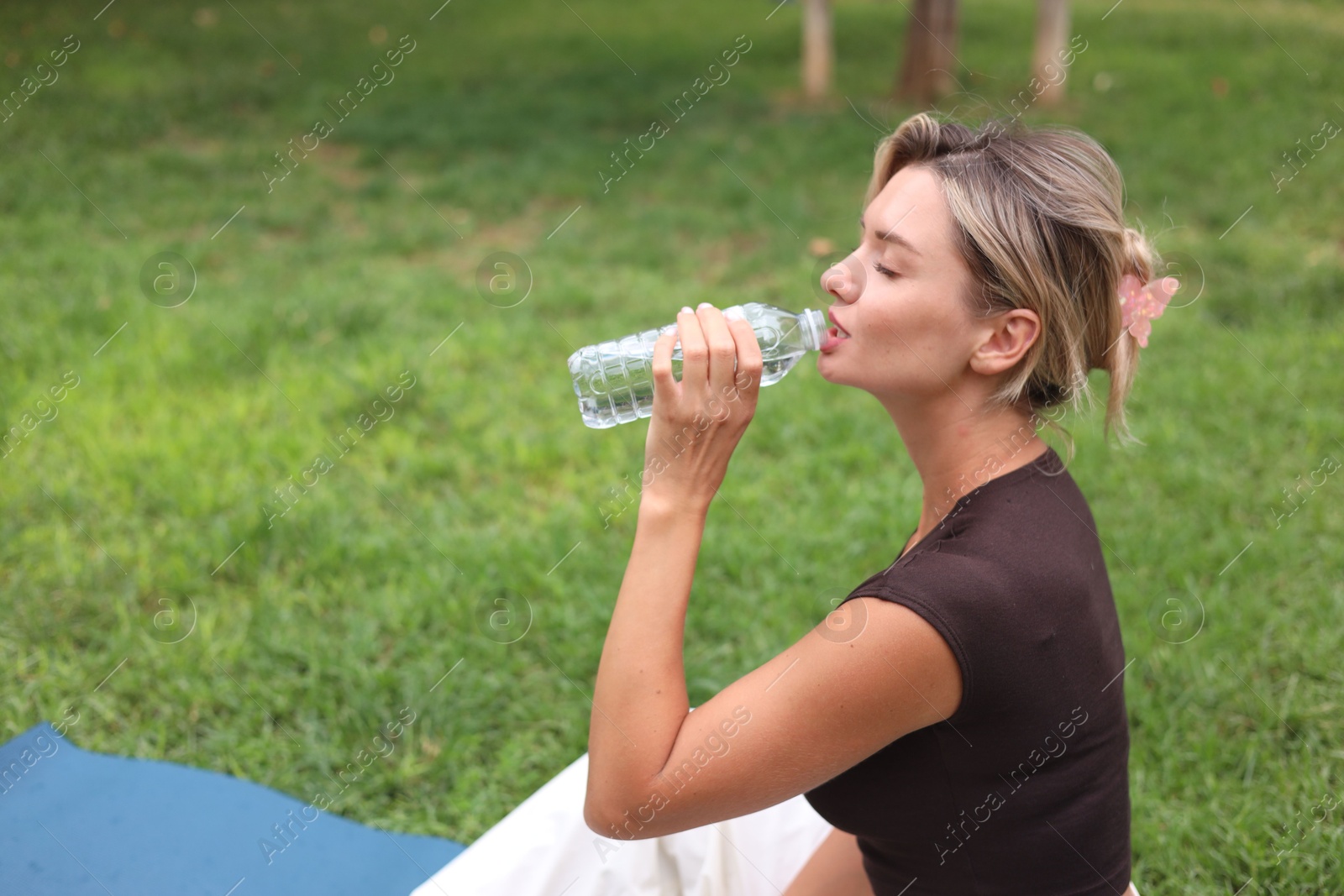 Photo of Young woman drinking water on yoga mat in park