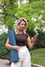 Photo of Beautiful young woman with yoga mat and phone outdoors