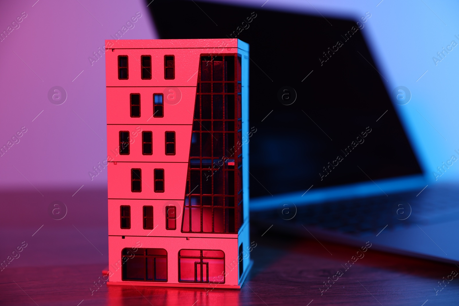 Photo of Real estate. Apartment building model and laptop on table in neon lights, closeup